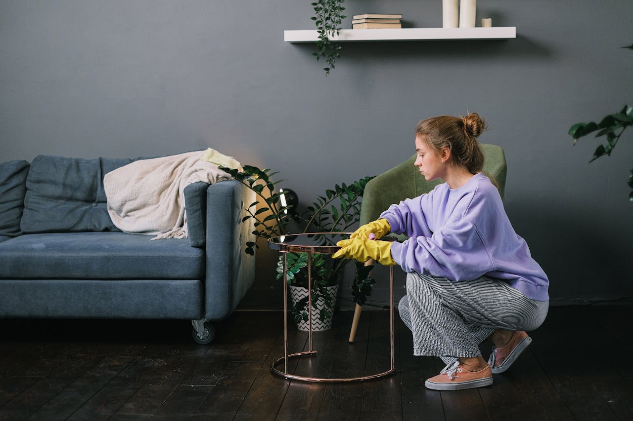 Side view of young female in latex gloves squatting down and wiping table in stylish living room in daytime