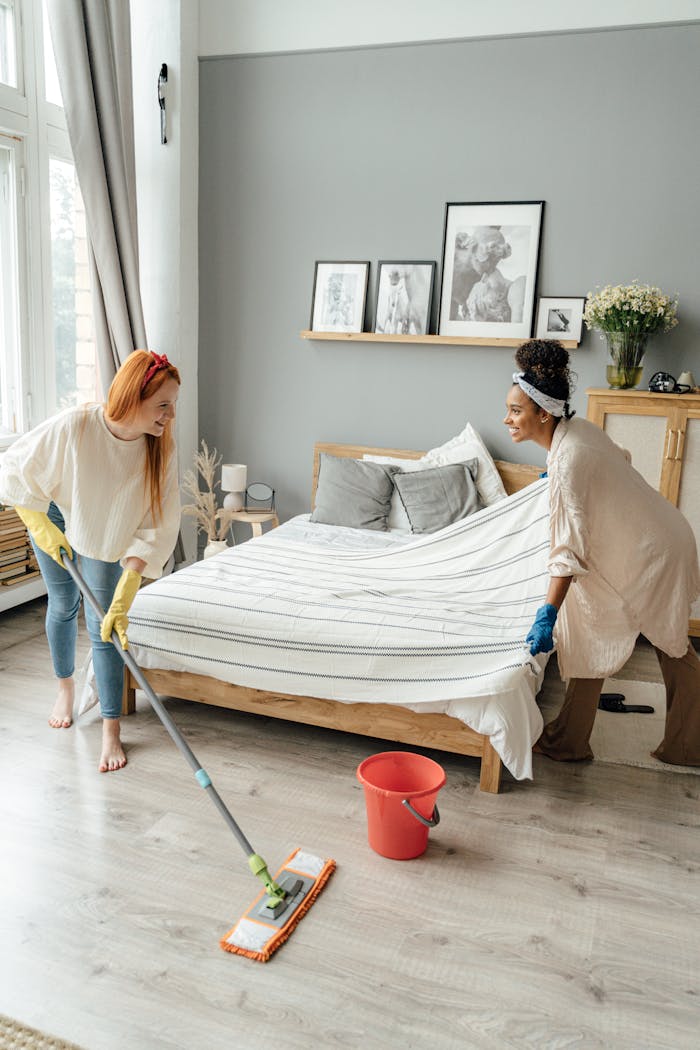 Two women enjoy house cleaning, tidying beds and floors in a bright bedroom.